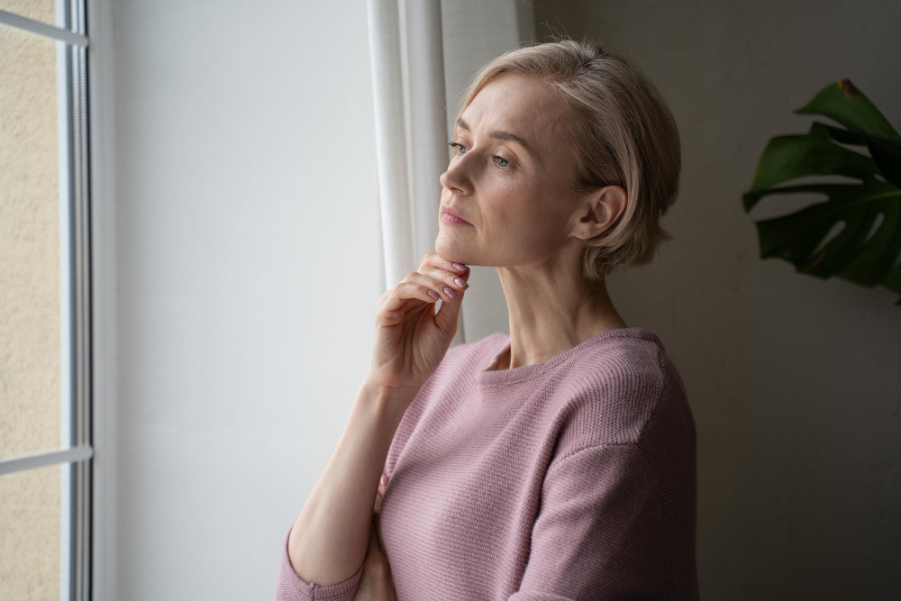 Mujer con jersey rosa mirando por una ventana, con una mano apoyada en la barbilla.